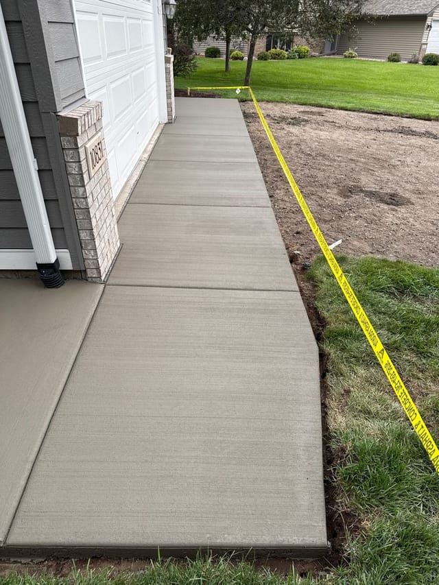 Newly poured concrete apron alongside a residential garage, bordered by yellow caution tape to prevent foot traffic while the concrete cures.