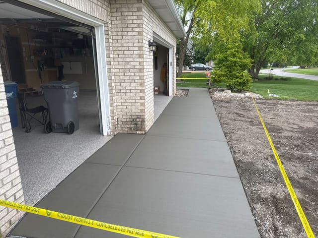 Newly poured concrete apron running alongside a brick house and open garage, marked off with yellow caution tape while curing.