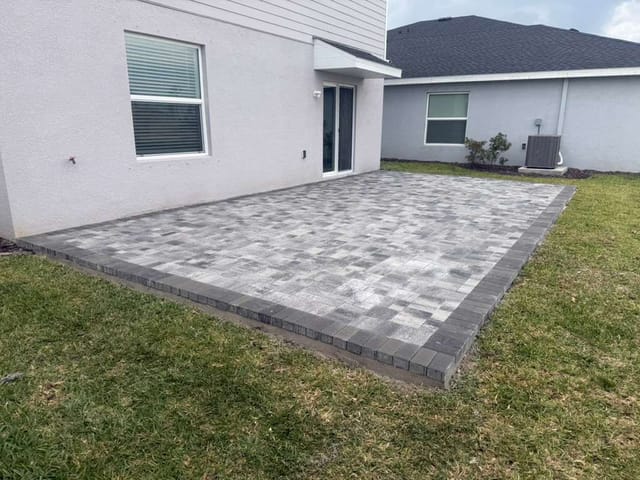 Large gray paver patio with a dark border beside a white house, surrounded by a grassy backyard.