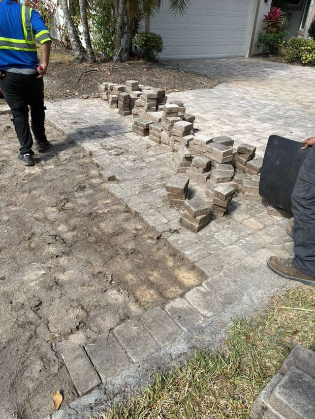 Driveway paver repair in progress, with stacked bricks on a partially removed section and workers standing nearby.