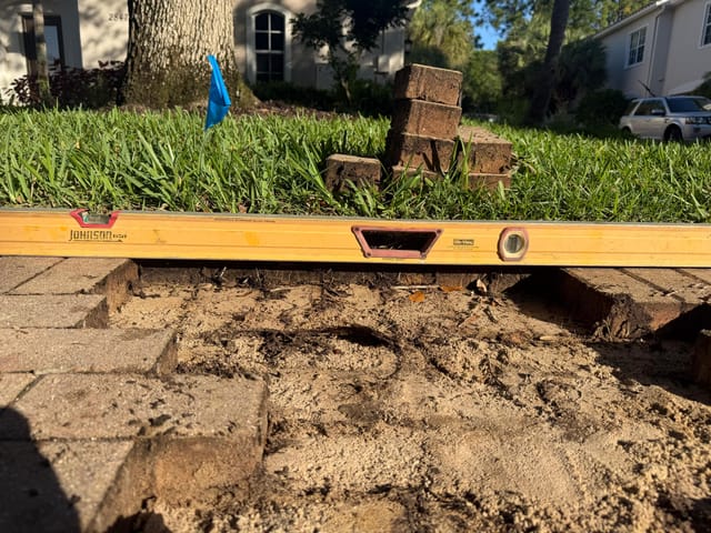 Close-up of a paver repair area with bricks removed, exposed sand base, stacked pavers, and a yellow level checking the surface.