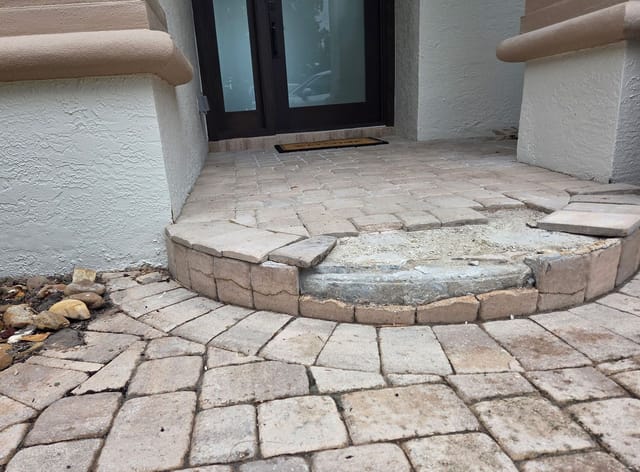 Front entryway with worn stone pavers, an uneven step, exposed concrete, and white stucco walls around dark glass doors.