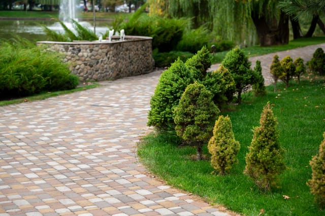 Landscaped paver sidewalk, trimmed evergreen shrubs, green grass, and a stone fountain near a pond in the background.