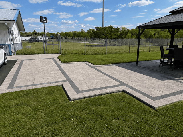 Outdoor patio with light pavers, dark accent borders, green lawn, chain-link fence, and a covered seating area under a blue sky.