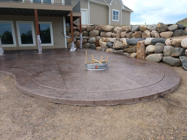 Large stamped concrete patio with a curved edge and built-in fire pit ring in the center, located beneath an elevated backyard deck.
