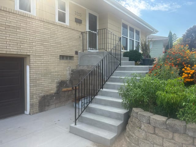 Newly poured concrete front steps and landing with black metal railings leading to the entrance of a brick home. The staircase rises from a concrete driveway, bordered by a landscaped flower bed with bright orange blooms and a retaining wall on the right.