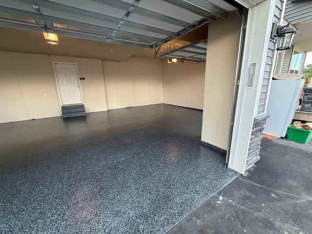 Open residential garage with a freshly finished black and gray speckled floor coating that extends wall to wall and slightly onto the driveway apron