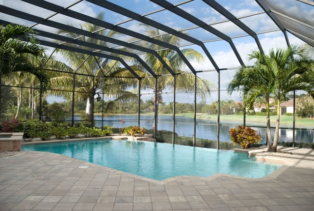 Screen-enclosed backyard pool with light stone tile deck, tropical landscaping, and views of a lake and palm trees through the mesh structure under a partly cloudy sky.