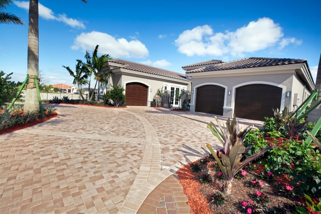 Modern single-story home with a wide paver driveway leading to three dark wooden garage doors, surrounded by tropical landscaping, palm trees, and bright flower beds under a blue sky with scattered clouds.