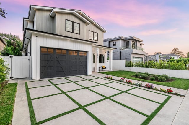 Modern farmhouse-style two-story home with a gray and white exterior, large dark wood garage doors, and a grid-patterned concrete driveway with artificial turf accents, surrounded by a manicured lawn and flower beds at sunset.