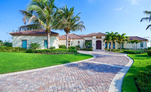 Single-story Mediterranean-style home with a red tile roof, tall arched wooden front door, and a curved paver driveway lined with manicured green grass and palm trees under a clear blue sky