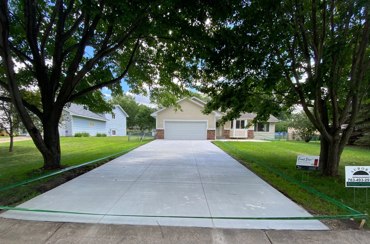 Finished concrete driveway replacement leading to a yellow house, bordered by lawn and mature trees.