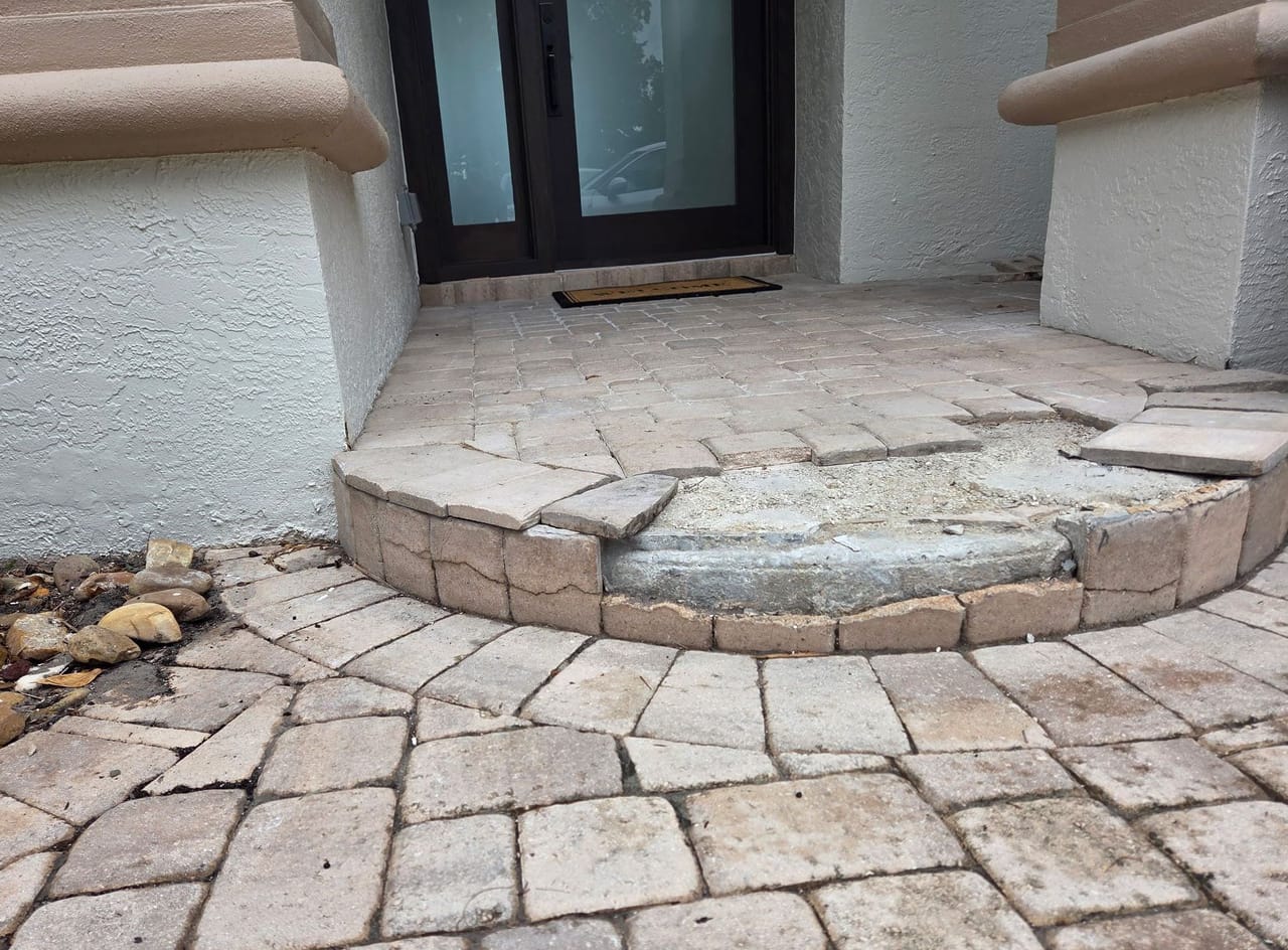 Front entryway with worn stone pavers, an uneven step, exposed concrete, and white stucco walls around dark glass doors.