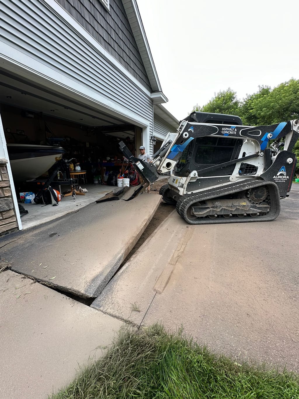 Heaved, cracked slab at a garage entrance before concrete driveway repair, with a track loader on site.