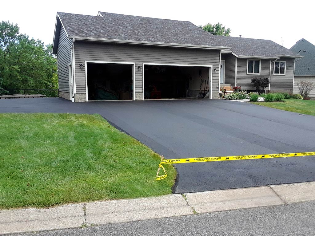 Newly paved asphalt driveway in front of a gray house with a two-car garage, green lawn, and yellow caution tape at the driveway entrance Maple Grove, MN.