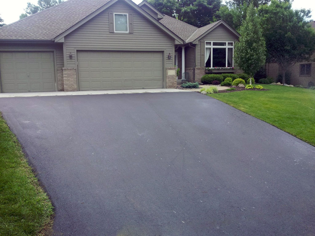 Newly paved asphalt driveway leading to a gray single-story house with a three-car garage, green lawn, and landscaped garden in Elk River, MN.